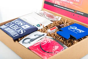 Gift box showing various contents for inspiration such as a mask, can cozy, door opener and a shirt